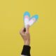 a person holds a heart with the colors of the transgender flag, blue, pink and white, in front of a yellow background with some blank space around