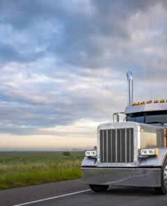 Gray classic big rig American semi truck with turned on lights and chrome exhaust pipes transporting covered cargo on flat bed semi trailer driving on the twilight highway road with stormy clouds sky