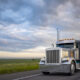 Gray classic big rig American semi truck with turned on lights and chrome exhaust pipes transporting covered cargo on flat bed semi trailer driving on the twilight highway road with stormy clouds sky